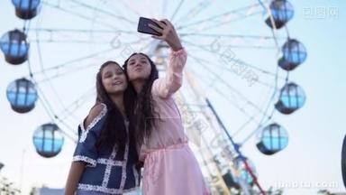 两个年轻女孩做自拍使用智能手机, 而站在一个摩天轮的背景。慢动作.